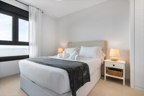 Ein ordentlich bezogenes Bett mit weißen Laken, einer grauen Decke und zwei Lampen auf einem Nachttisch mit einem gewebten Korb.