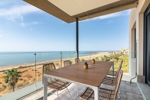 Ein Balkon mit Holztisch und Korbstühlen, der auf einen Strand und das Meer blickt.