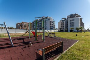 Ein Spielplatz mit Schaukeln und Rutsche, umgeben von einem befestigten Bereich und Gebäuden im Hintergrund.