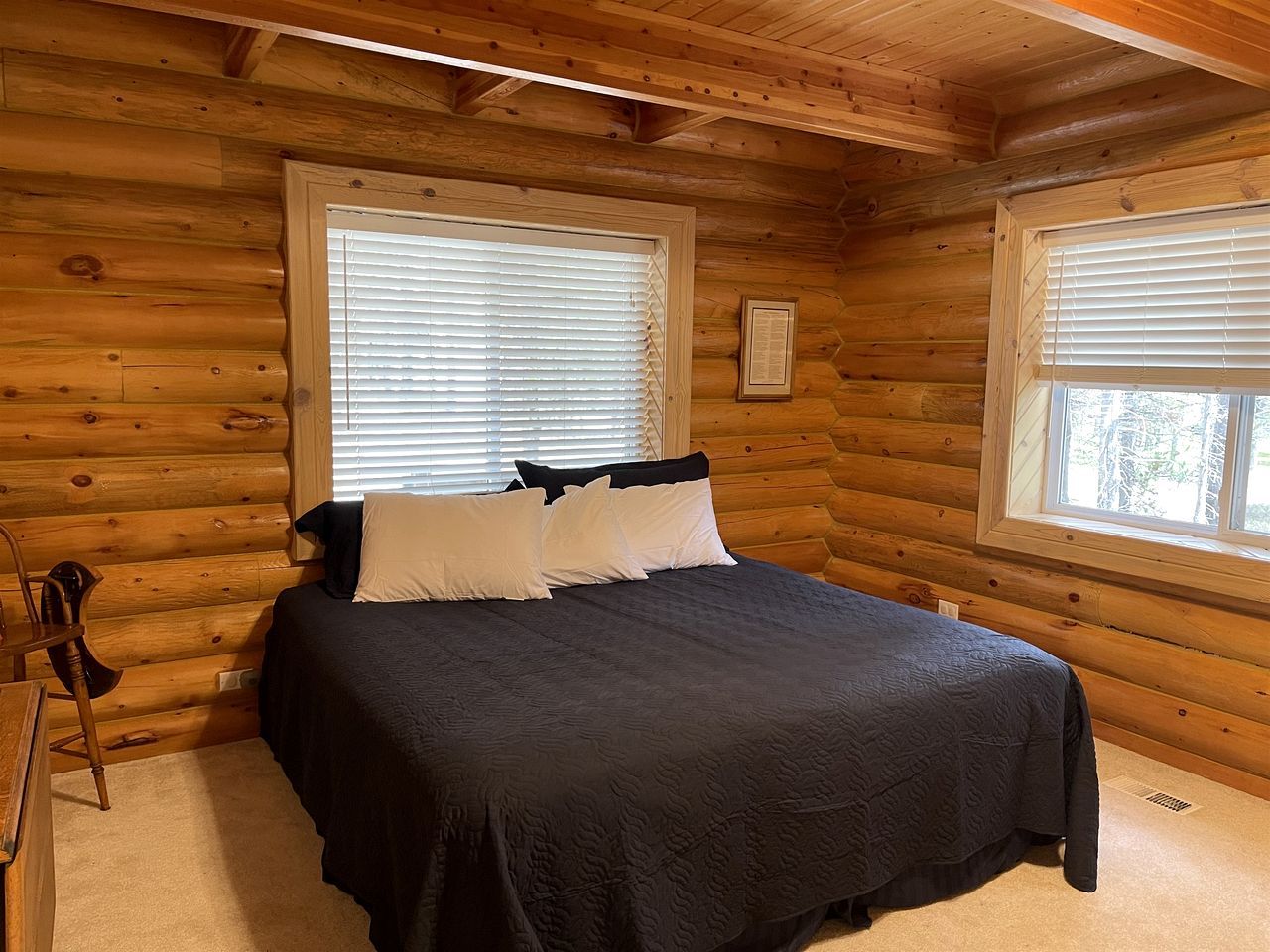 A log cabin bedroom with a bed, two windows, a framed picture, and a chair.