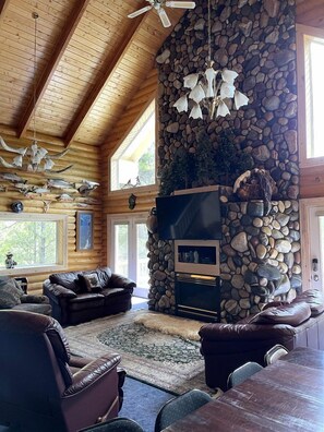 A log cabin interior with a stone fireplace, leather furniture, and a ceiling fan. - Cozy Log Cabin Close to Yellowstone National Park, Idaho (Island Park)