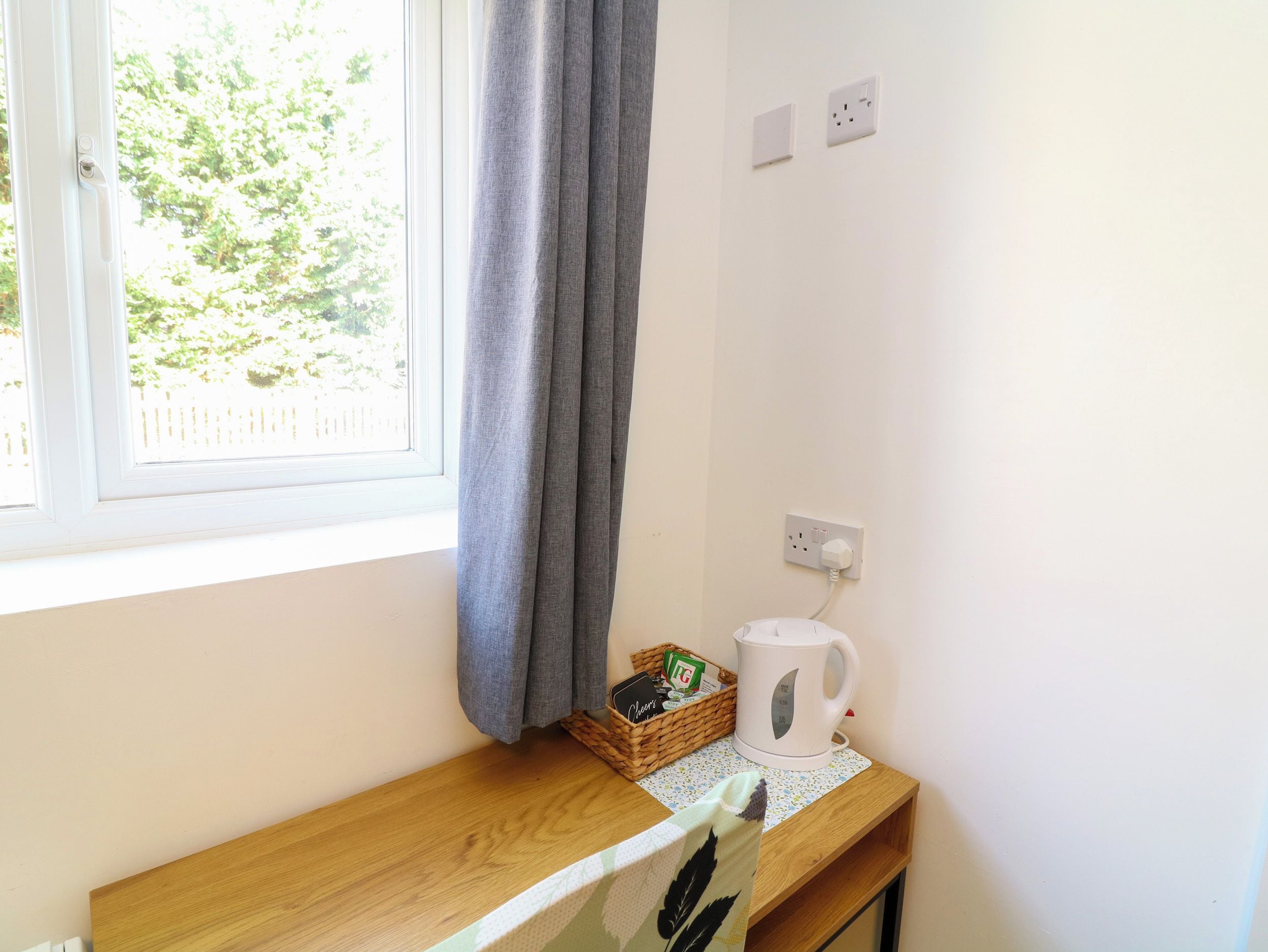 A room with a window, a wooden table, a kettle, a basket, and a power outlet.