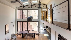 Un ambiente spazioso con soffitto alto, lucernari, zona pranzo con un tavolo lungo e sedie, e una scala che conduce a un livello superiore.