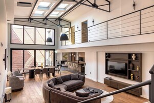 A spacious living area with a large sofa, dining table, and chairs, a kitchen in the background, and a TV mounted on the wall.