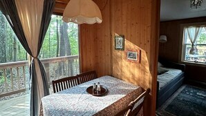 A wooden cabin interior with a dining table, chairs, a hanging light fixture, and a bed with a view of the outdoors.