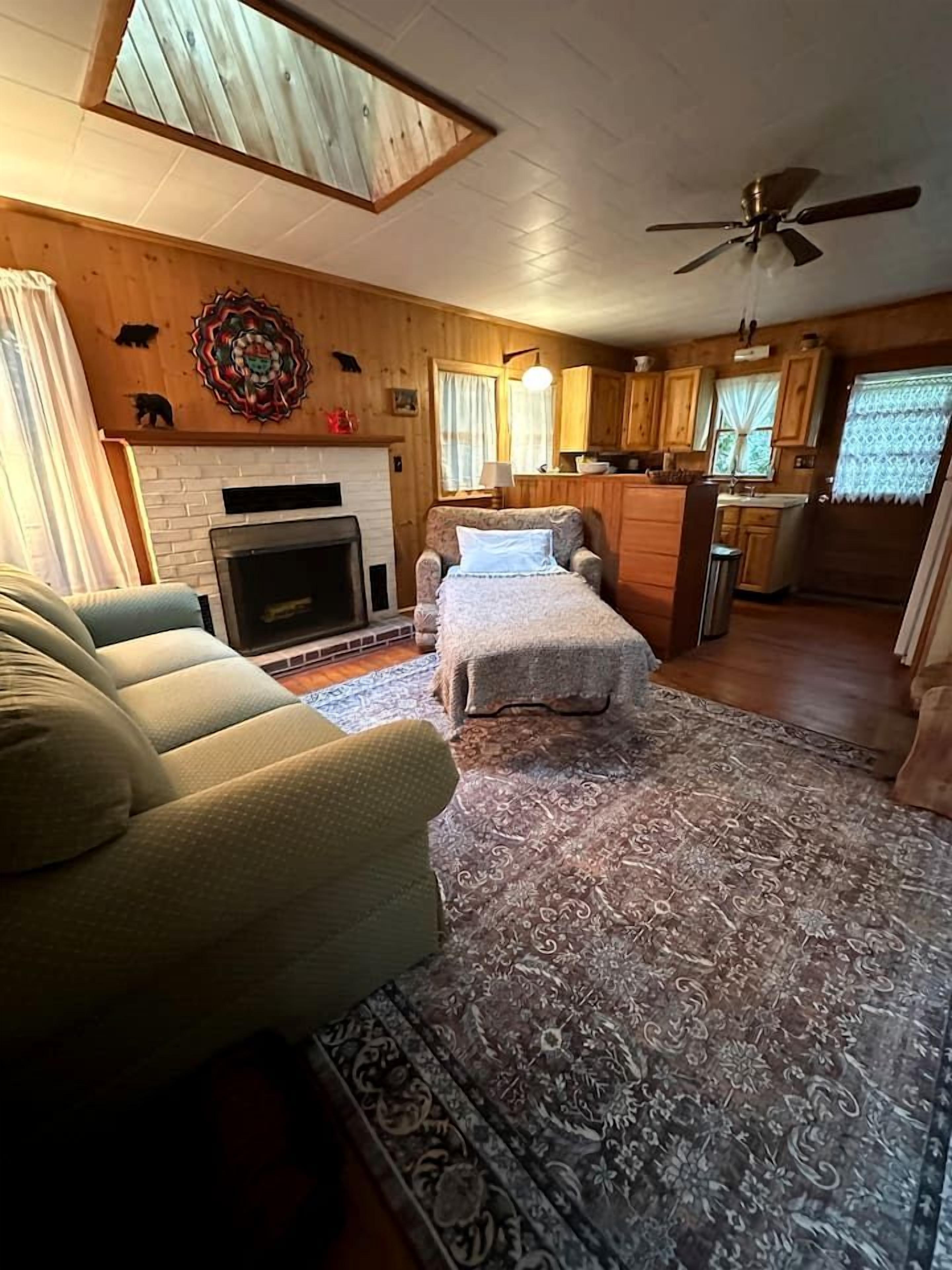 A cozy living room with a fireplace, a sofa, a bed, and a kitchen area with wooden cabinets and a ceiling fan.