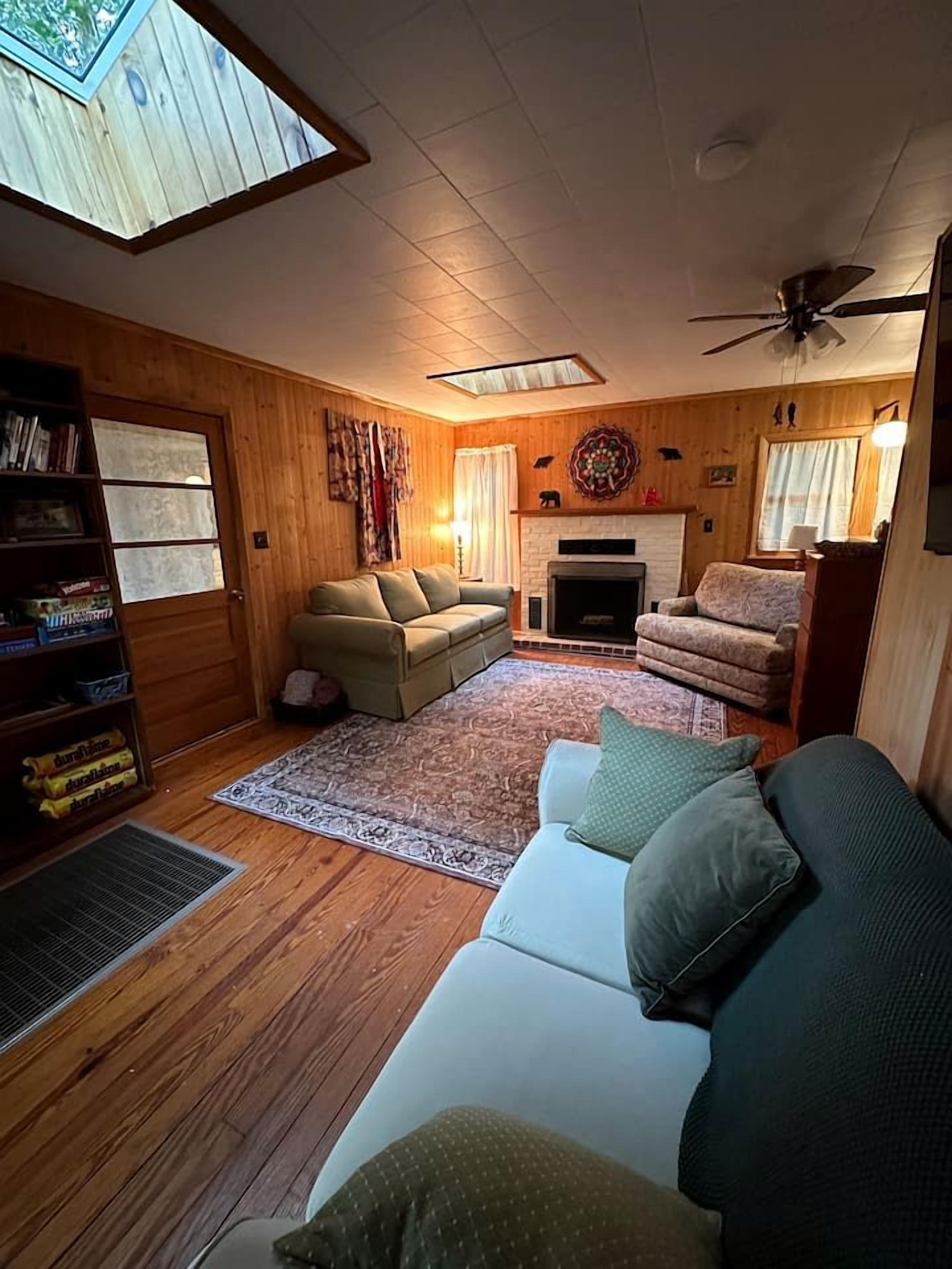 A cozy living room with a fireplace, wooden walls, a ceiling fan, and a skylight.