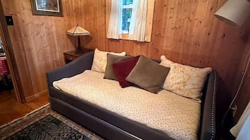 Ein Zimmer mit holzverkleideten Wänden, einem Bett mit gemusterten Textilien, einem Fenster mit Vorhängen, einer Nachttischlampe und einem Teppich.
