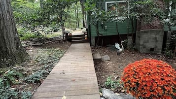 A wooden pathway leading to a house surrounded by trees and a red flowering bush.