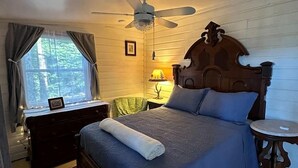 A bedroom with a bed, a ceiling fan, a nightstand, a lamp, a window with curtains, and a small table.