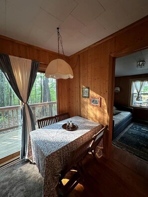 A wooden cabin interior with a dining table, chairs, a hanging light fixture, and a bed with a view of the outdoors. - Hidden Countryside Gem with Fire Pit & Tranquil Setting in Aspers, Pennsylvania (Aspers)