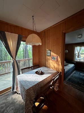 Interior de una cabaña de madera con mesa de comedor, sillas, lámpara colgante y una cama con vistas al exterior.
