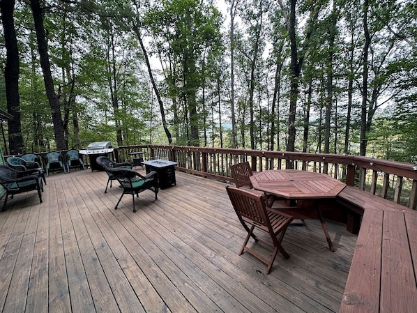Una terraza de madera con mobiliario exterior, una parrilla y vistas al bosque.