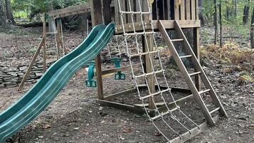 Eine Holzspielanlage mit Rutsche und Kletternetz in einem bewaldeten Gebiet.