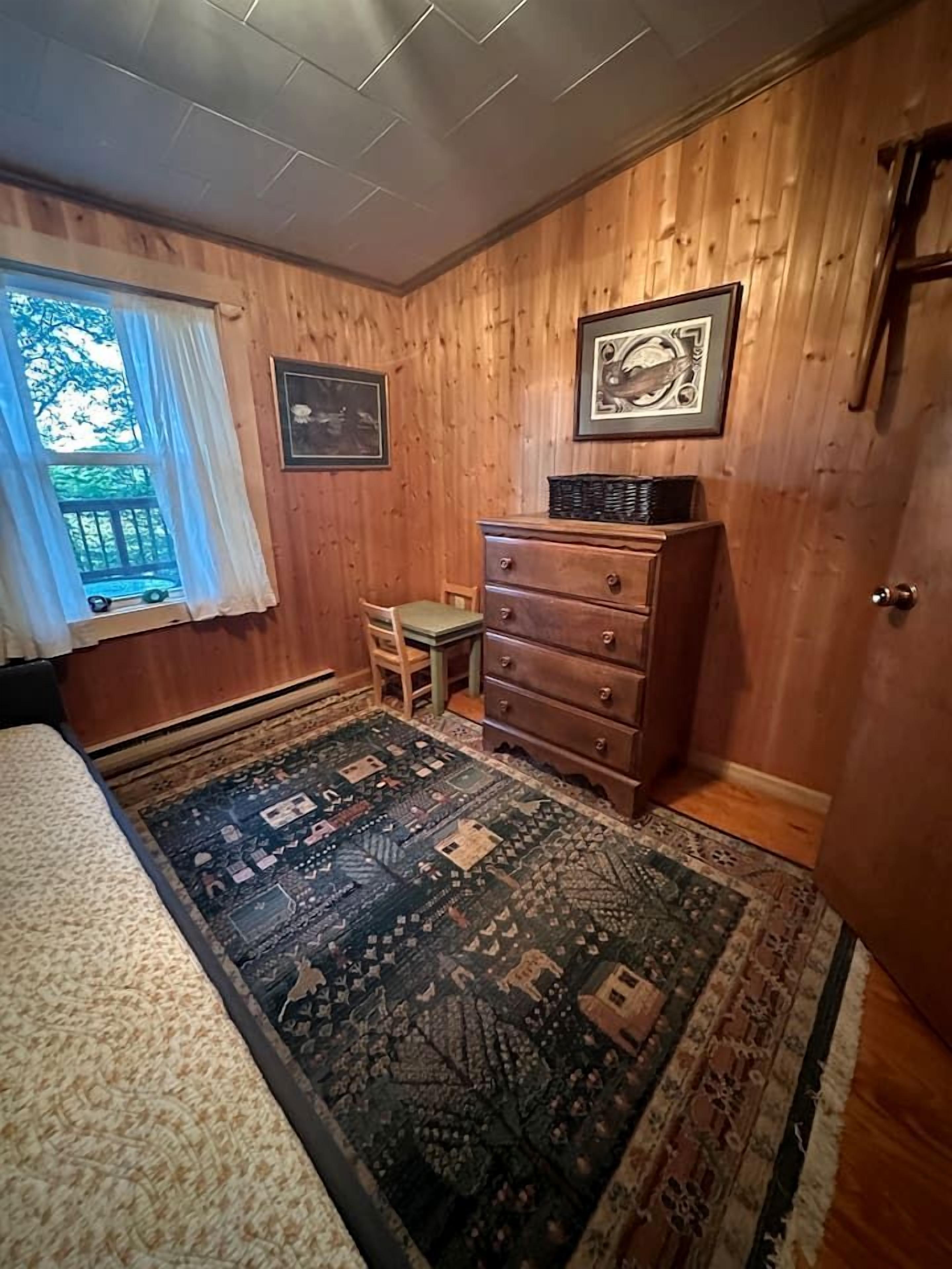 A room with wooden paneled walls, a bed, a dresser, a chair, and a rug.