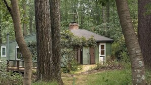 A small cabin with a chimney, surrounded by trees and a wooden deck.