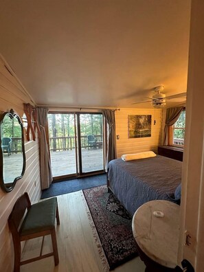 A bedroom with a bed, a chair, a small table, a mirror, and a sliding glass door leading to an outdoor deck.