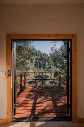 Puerta abierta que da a un balcón con mesa y sillas, con vistas a una ladera boscosa.
