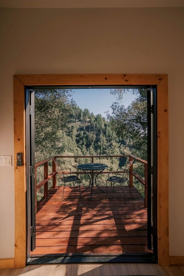 Puerta abierta que da a un balcón con mesa y sillas, con vistas a una ladera boscosa.