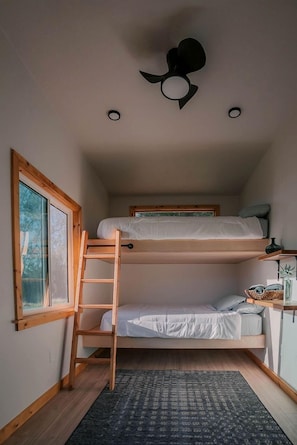 A compact room with a bunk bed, a ladder, a desk with a chair, and a window with a view of trees.