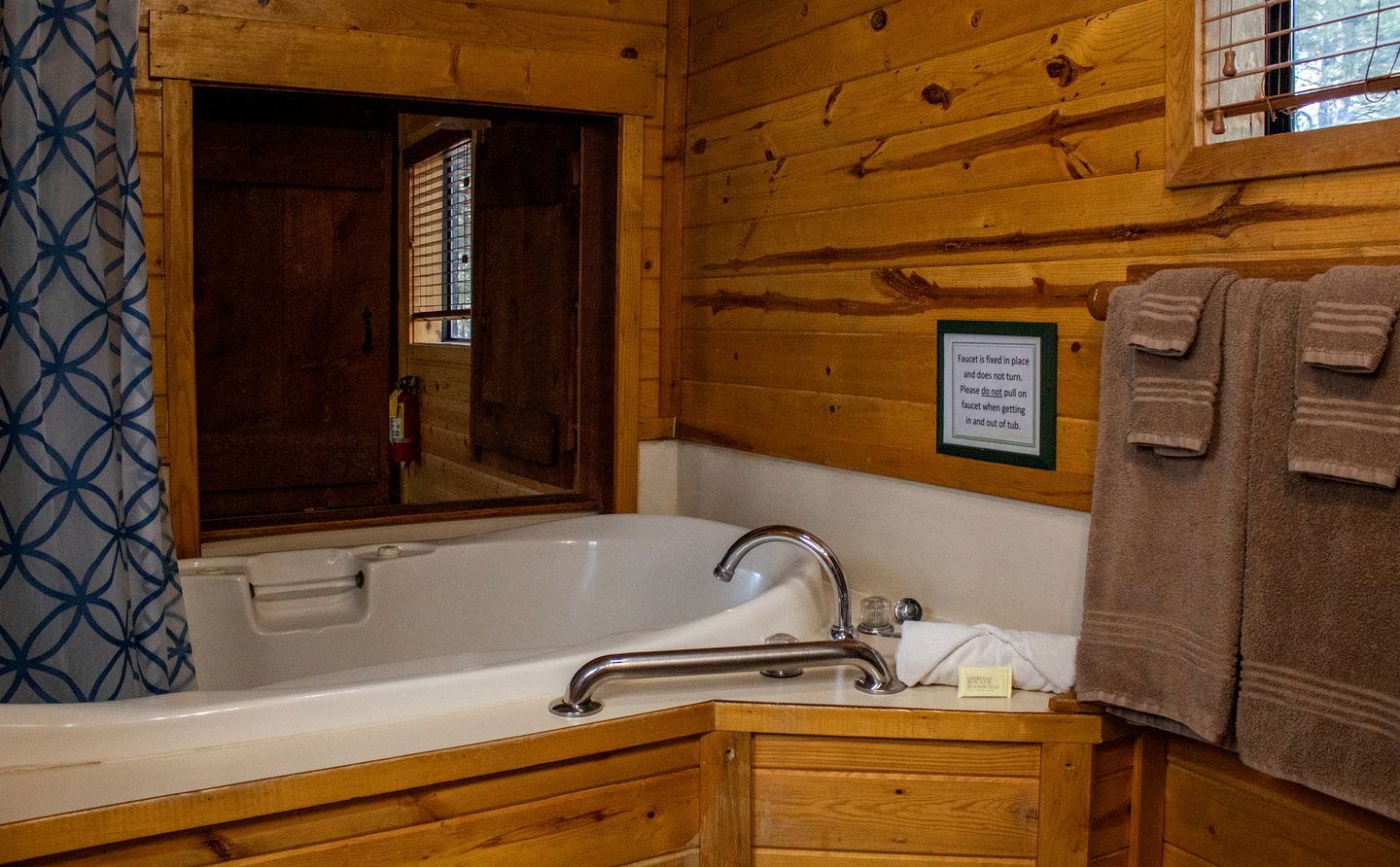 A wooden cabin bathroom with a bathtub, a towel rack, and a window with blinds.