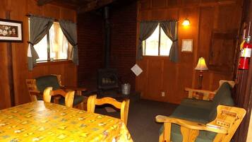 Une pièce chaleureuse avec un décor intérieur en bois, une table recouverte d’une nappe jaune, deux chaises, une cheminée et une lampe.