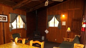 Une pièce chaleureuse avec un décor intérieur en bois, une table recouverte d’une nappe jaune, deux chaises, une cheminée et une lampe.