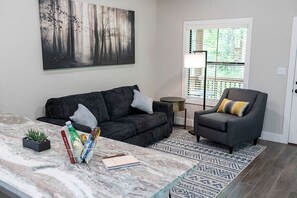 Ein Wohnzimmer mit einem dunkelgrauen Sofa, einem grauen Sessel, einem Marmortisch, einer kleinen Pflanze und einem Bild von einem Wald.