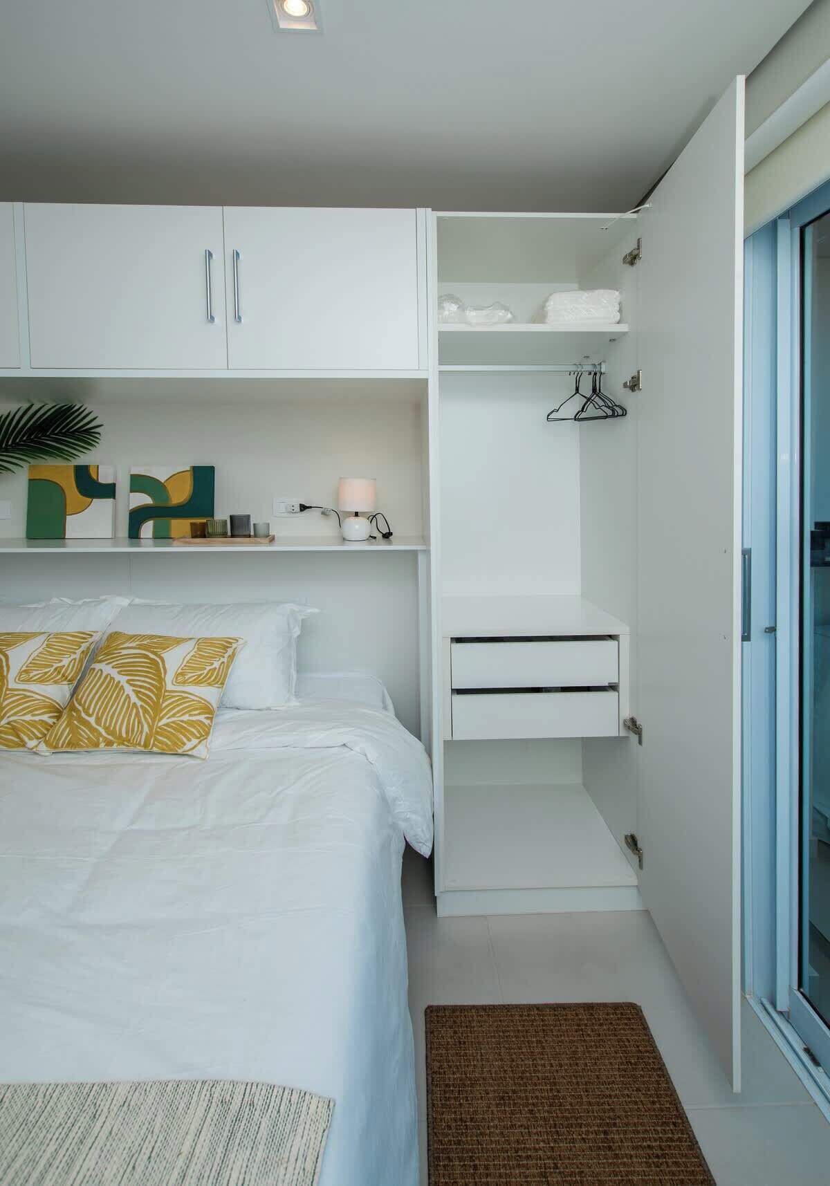Quarto com cama, armários brancos, mesa de cabeceira, luminária pendente e porta para uma varanda.