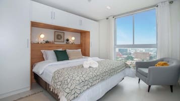 A bedroom with a bed, a chair, a large window, and a view of buildings.