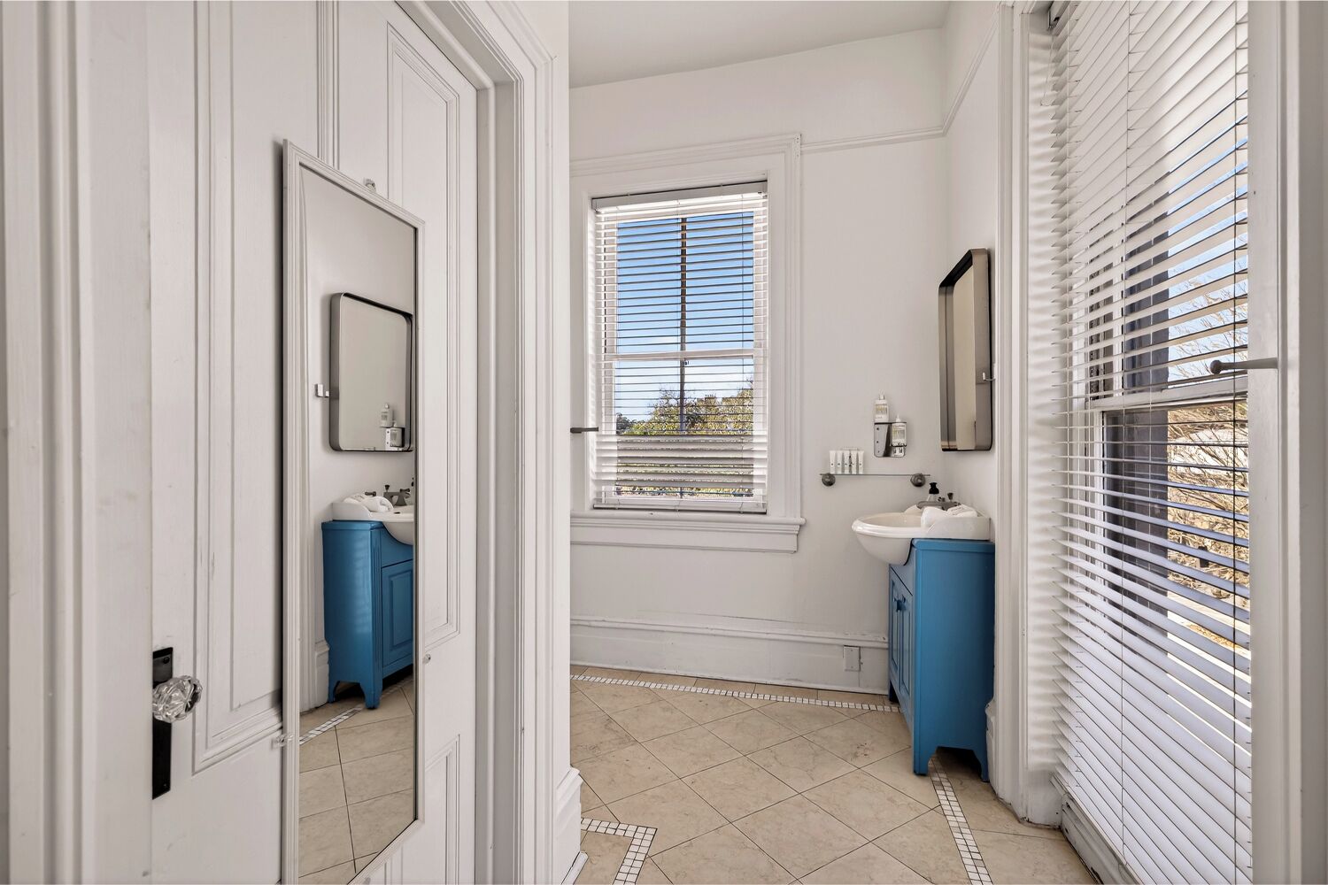 A bathroom with white walls, a window with blinds, two sinks with blue cabinets, and a mirror.
