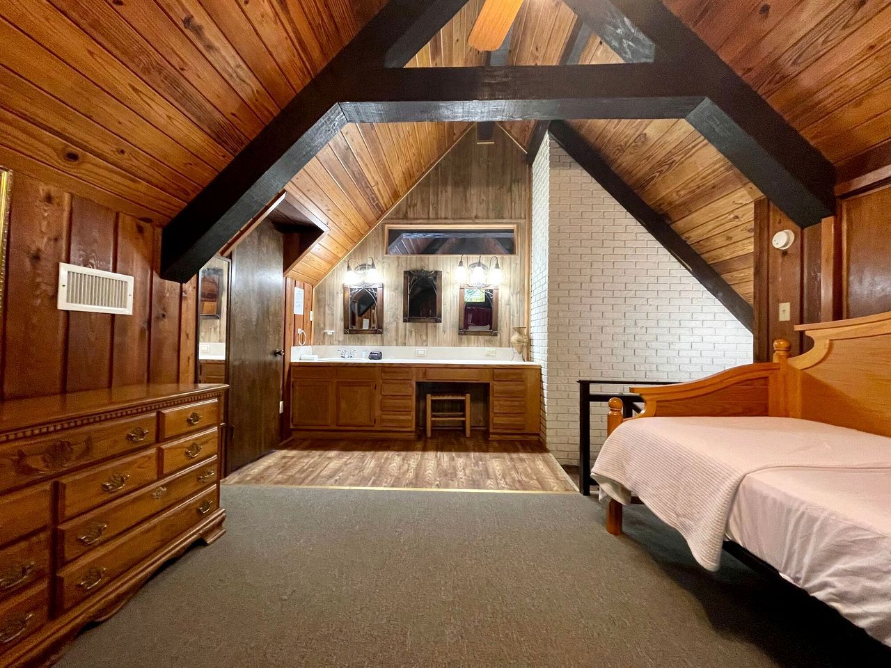 A bedroom with a wooden ceiling, a bed, a dresser, a vanity with a mirror, and a small stool.