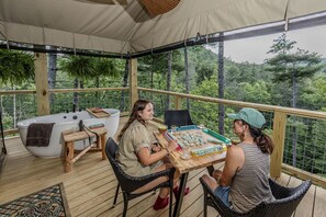 Une terrasse en bois avec un jacuzzi, une table pour jouer à des jeux de société et des chaises en osier.