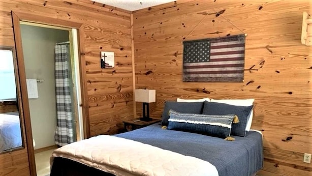 Un dormitorio con paneles de madera, con una cama, una mesita de noche, una lámpara, una bandera estadounidense y una puerta que conduce a un baño.