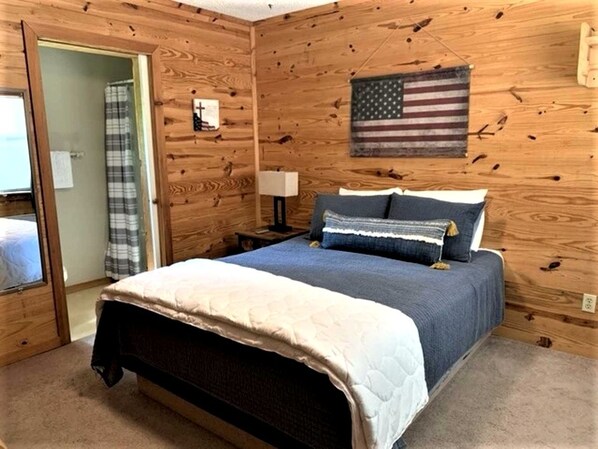 A wooden-paneled bedroom with a bed, a nightstand, a lamp, an American flag, and a door leading to a bathroom.
