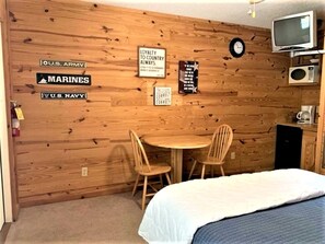 A room with a wooden wall, a bed, a desk with two chairs, and a television.