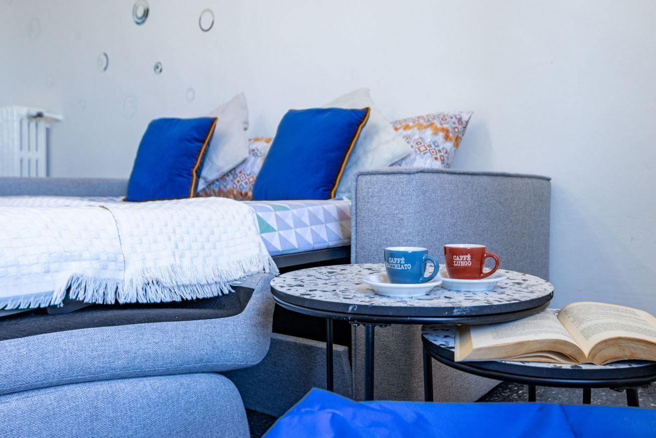 Ein Bett mit blauen und weißen Kissen, ein kleiner runder Tisch mit zwei Tassen und einem offenen Buch sowie ein grauer Sessel.