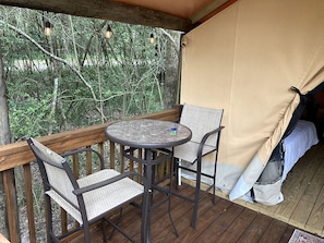 Um deck de madeira com uma mesa redonda e duas cadeiras, uma tenda e vista para árvores.