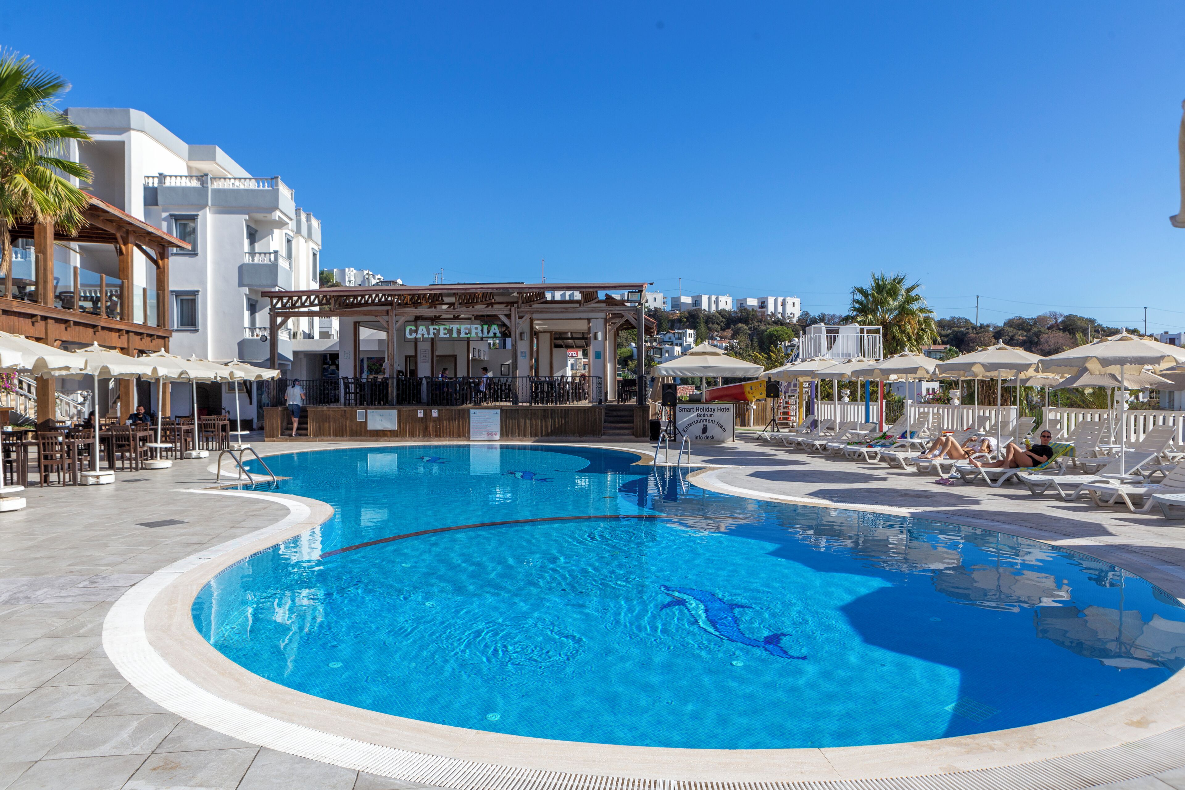 A swimming pool with lounge chairs, a cafe, and a building with a sign reading 'CAFETERIA'.