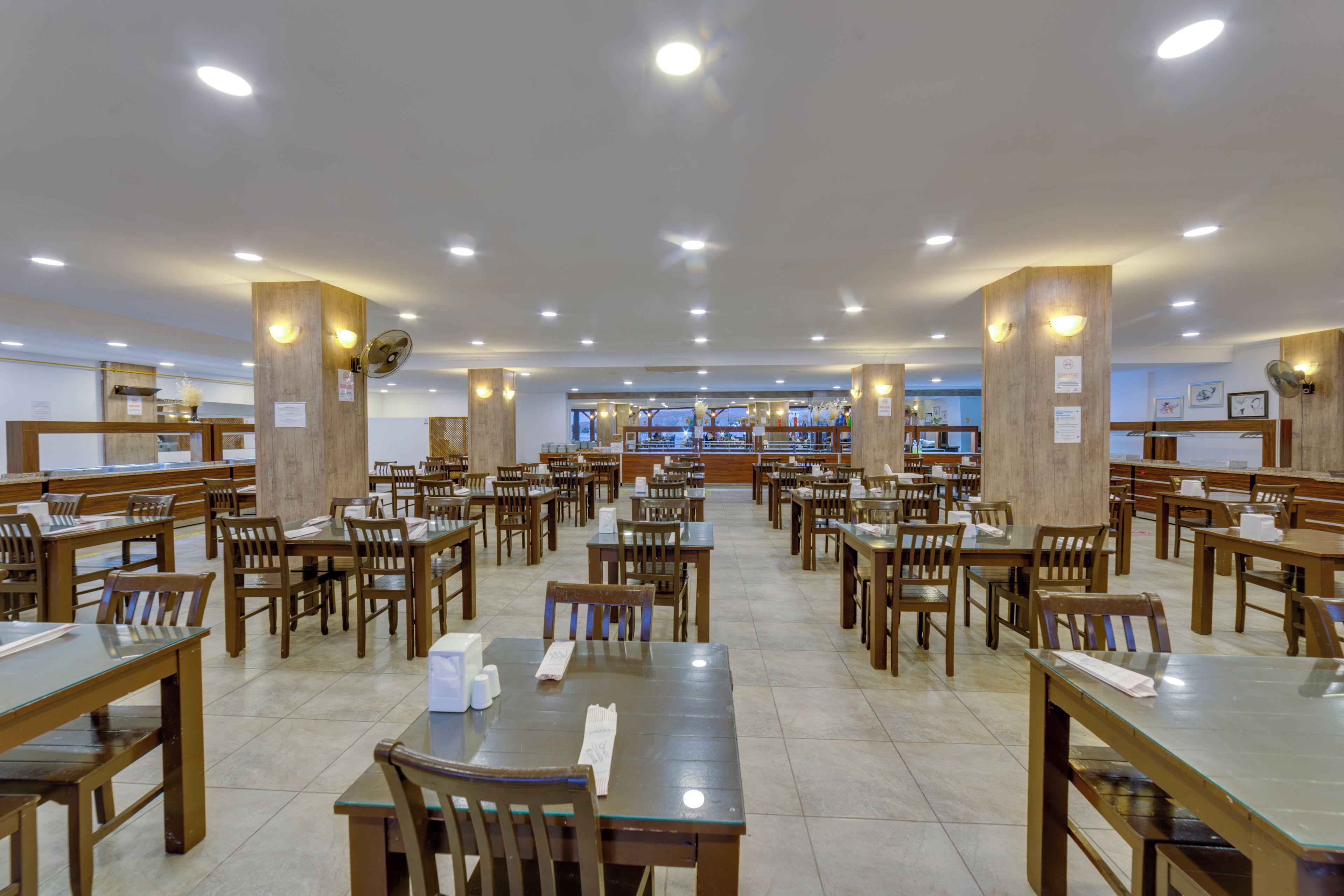 A spacious dining area with multiple wooden tables and chairs, a central counter, and ceiling lights.