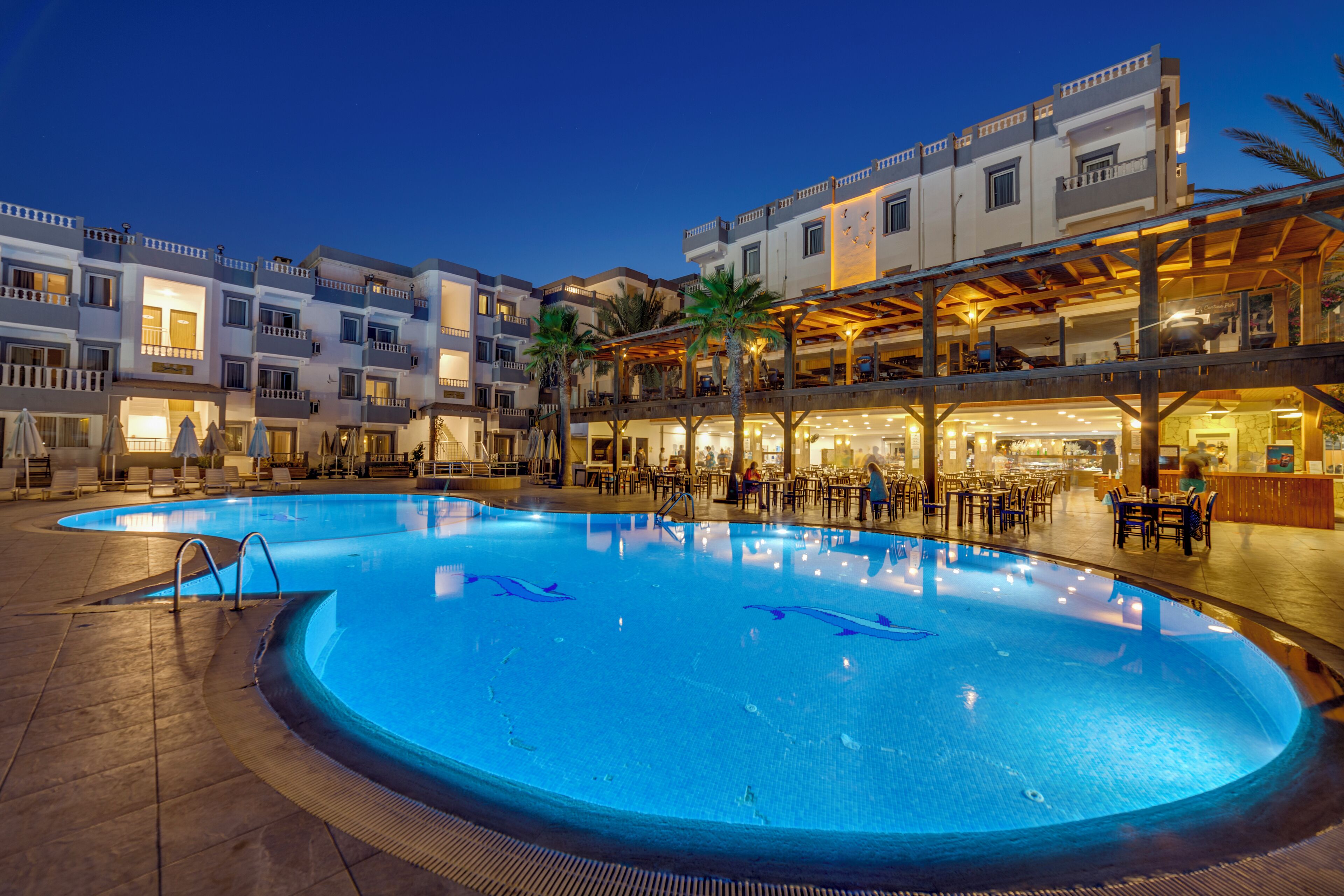 Una piscina con un área central hundida, rodeada de mobiliario exterior y una zona cubierta con mesas y sillas.