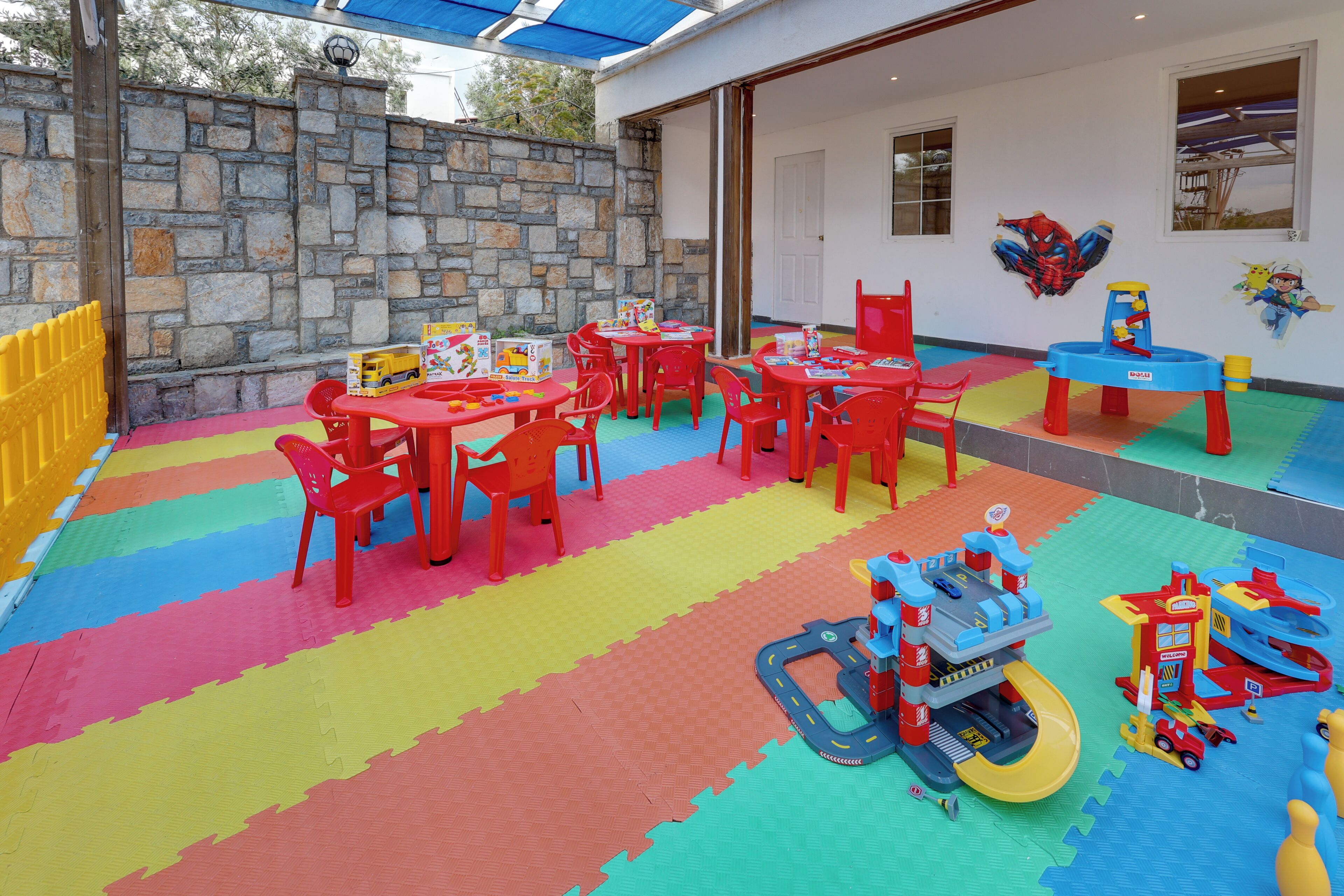 Zona de jocs infantils amb estores de colors, taules vermelles i elements de joc.
