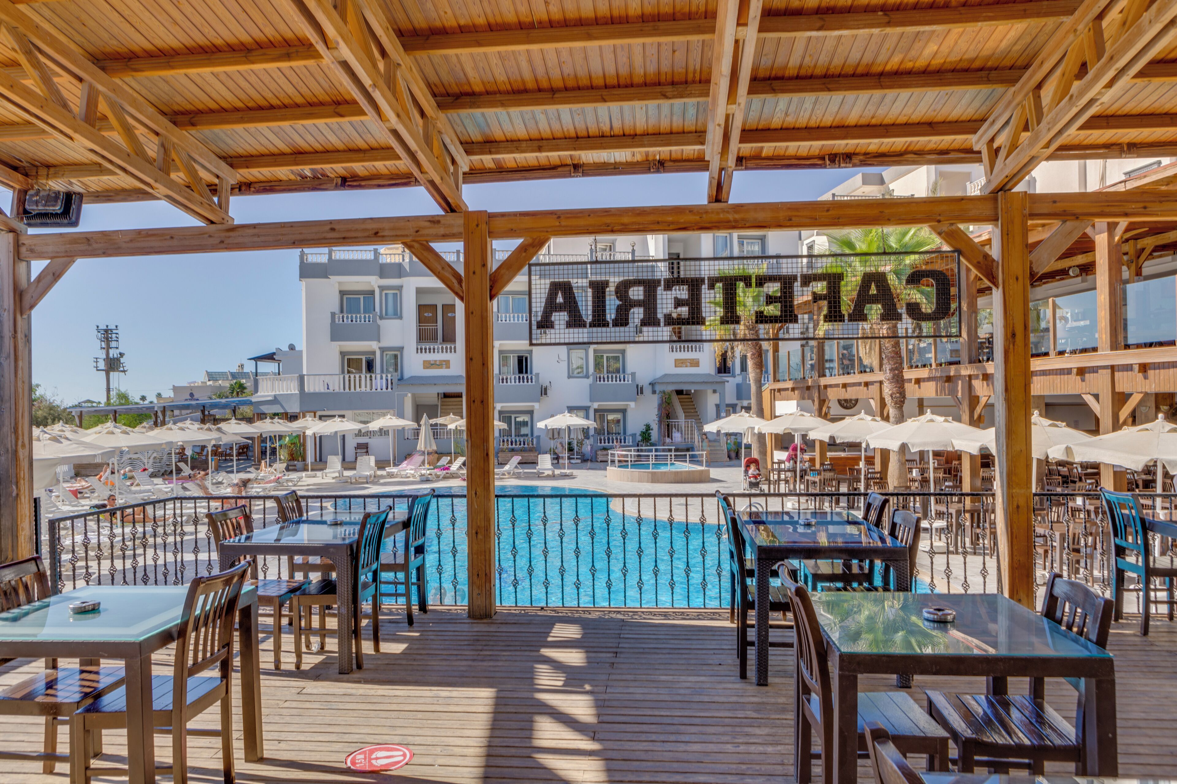 A wooden deck with tables and chairs, a pool, and a building with balconies in the background.