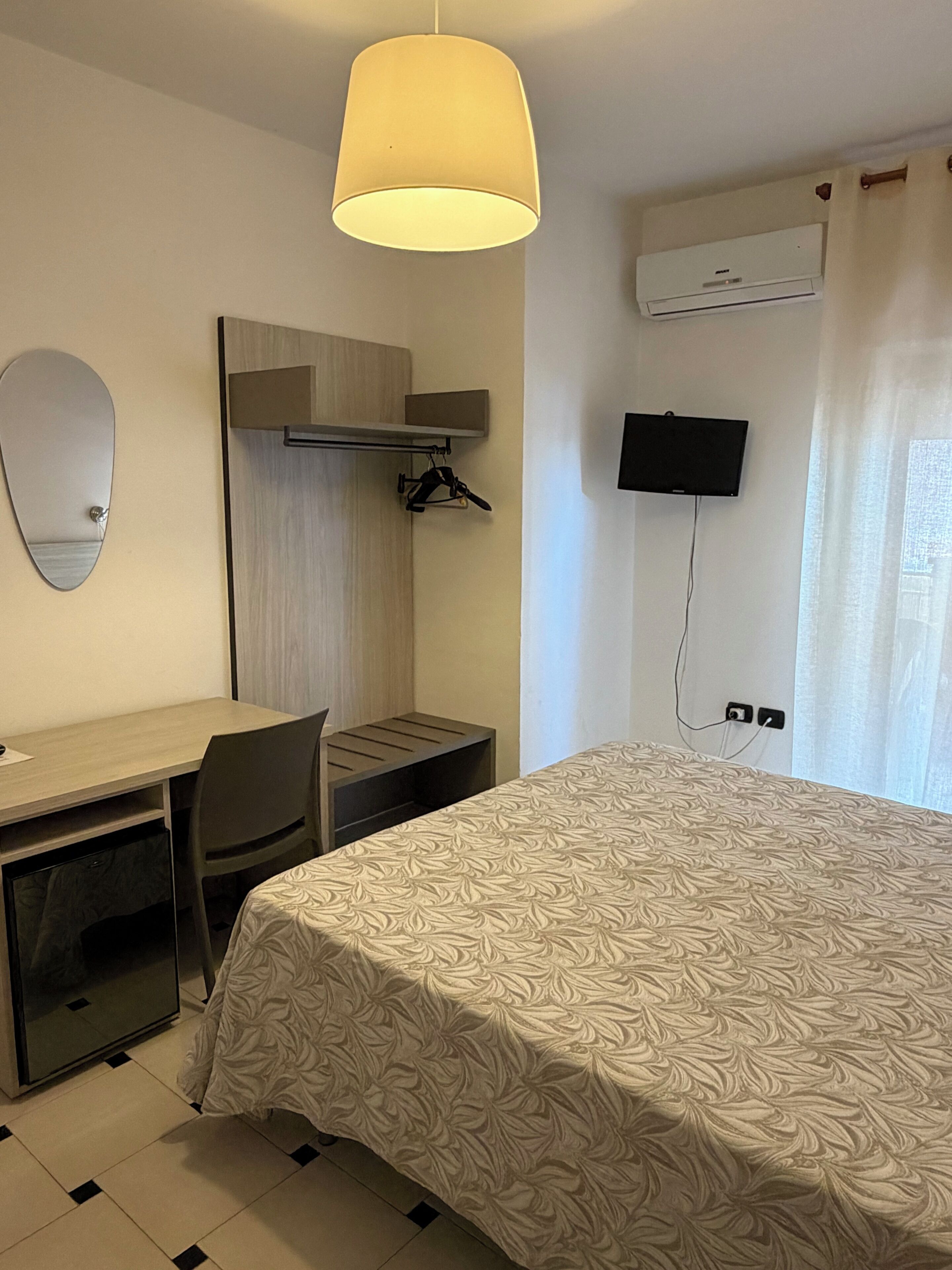 Habitación de hotel con cama, escritorio, silla, espejo, televisor y aire acondicionado.