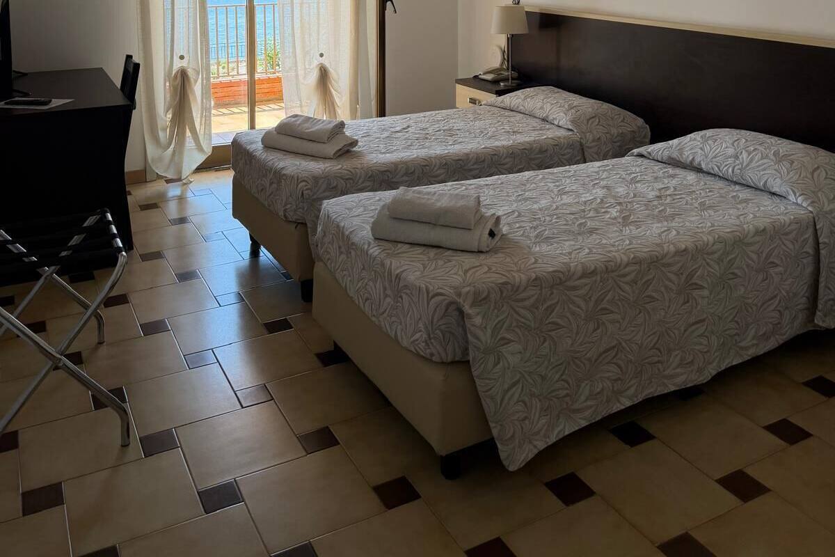 Ett hotellrum med två sängar, ett skrivbord, en stol, en takfläkt och utsikt över havet.