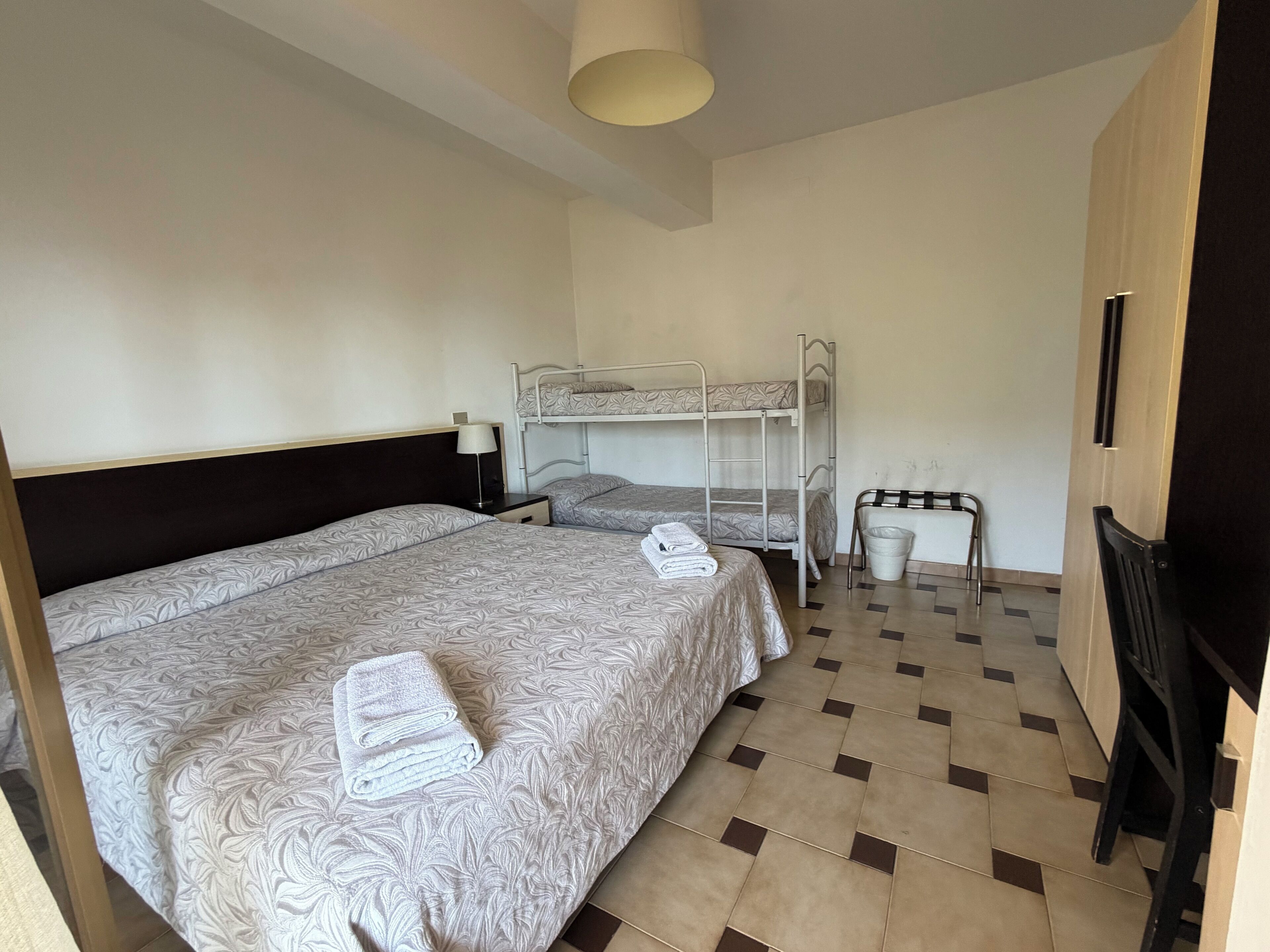 Une chambre avec un lit superposé, un lit simple, un bureau avec une chaise, une lampe et une poubelle.