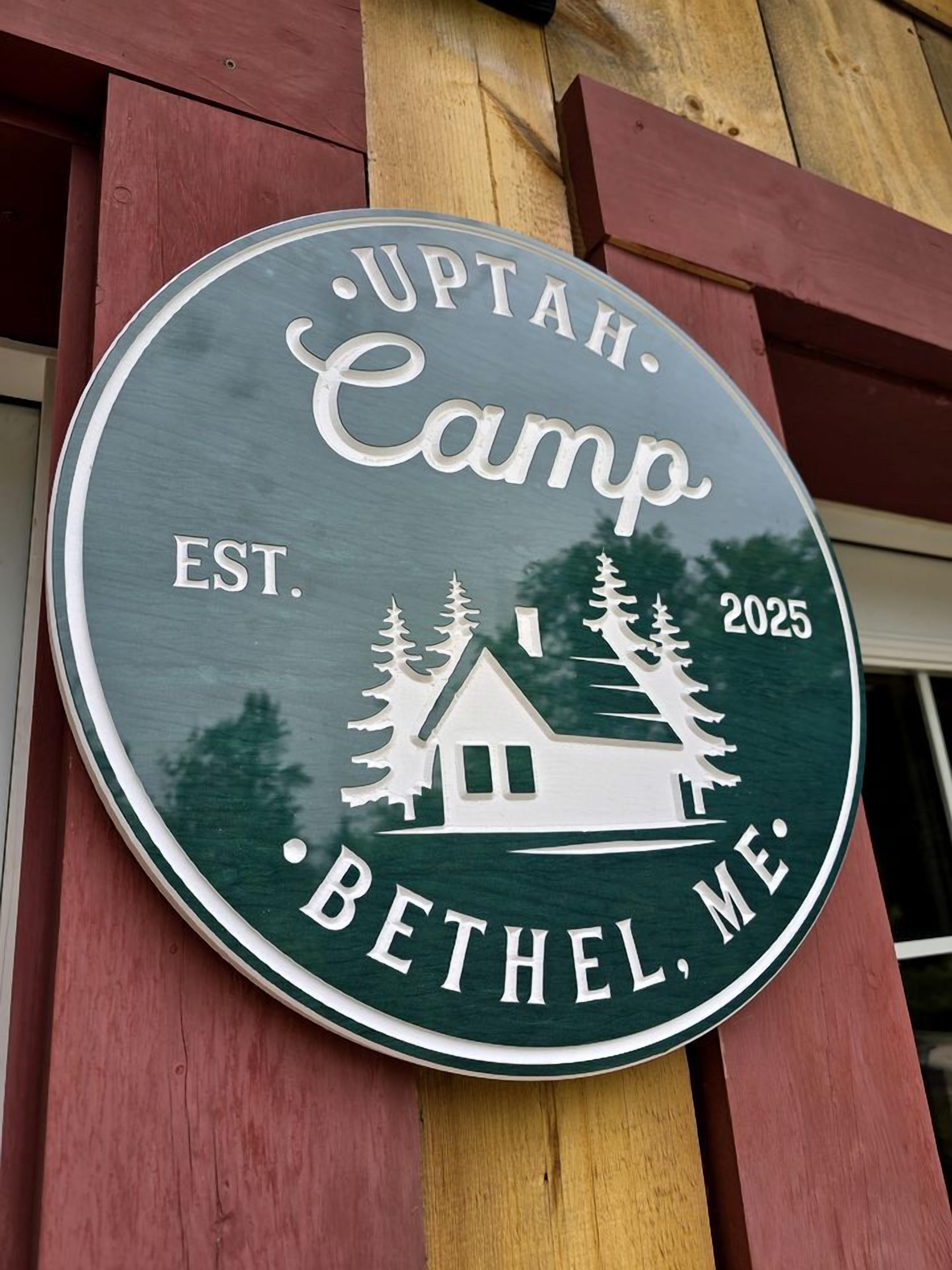 Segno circolare con il testo 'UpTah. Camp Bethel, ME. EST. 2025' e un'illustrazione di una baita.