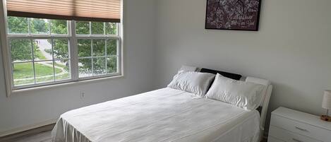 Un dormitorio con cama, mesita de noche, una obra de arte en la pared y una ventana con persianas.