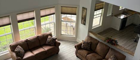A spacious living room with a brown leather sofa, a ceiling fan, and large windows offering a view of the outdoors.