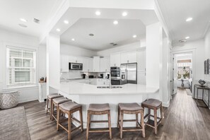 A modern kitchen with white cabinetry, a central island with bar stools, and a large window with blinds. - Le Destin Gulf Views Private Pool Private Beach (Miramar Beach)